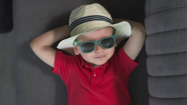 Cute toddler in sunglasses and hat lies on couch smiling