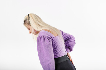 Side view of young blonde woman wearing lilac blouse isolated over white background suffering from pain in lower back. Feels back pain massaging aching muscles, sad suffers from low-back lumbar pain.