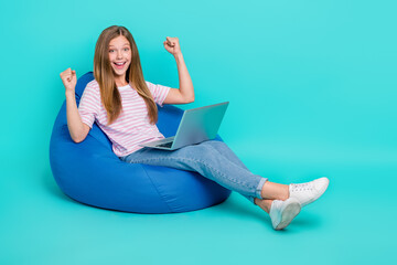 Full length photo of pretty lucky girl dressed pink t-shirt bean bag rising fists gadget empty space isolated teal color background