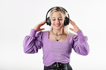 Young blonde girl smiling happy wearing lilac ribbed blouse isolated over white background closed eyes, listens to favorite music, touches headphones and enjoys it immensely.