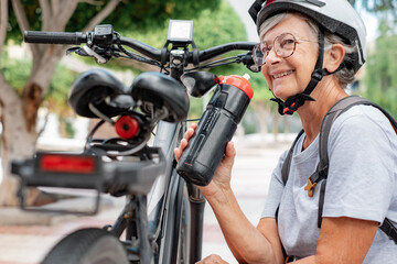 Obraz premium Smiling senior cyclist woman in urban park wearing helmet and backpack drinking from water bottle resting close her electro bicycle. Concept of healthy lifestyle and sustainable mobility