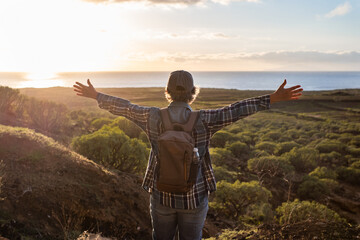 Back view of senior caucasian woman with hat and casual clothing standing with open arms looking at the horizon over sea. Elderly lady enjoying nature and freedom admiring landscape at sunset light