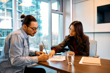 Asian financial advisor showing her client where to sign contract during meeting in office.