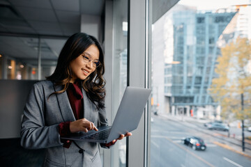 Happy Asian businesswoman working on laptop by window.
