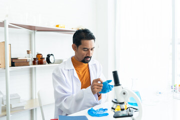 A male teacher in white lab coat wearing rubber gloves, preparing many laboratory tools shelves with microscope, textbooks, yellow and blue chemical flasks on table in university science classroom.