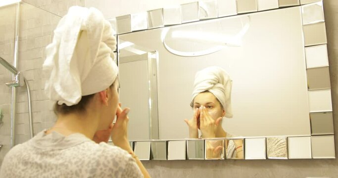 Fresh, nice female with bath towel on head looking at mirror reflection, applying white product moisturizing cream