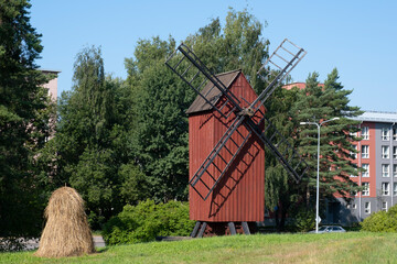 Old red wooden windmill with a haystack in the foreground. Modern apartment building in the background. Finnish agriculture.