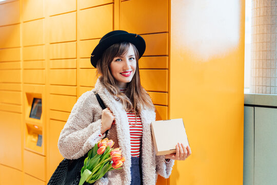 Portrait of smiling fashion woman with box near modern postal automatic mail terminal with self service device for pickup or refund an order. Electronic locker for storing parcels. Selective focus
