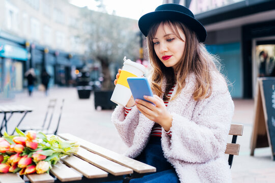 Stylish young woman enjoying coffee from reusable cup, reading text message on mobile phone at coffee shop . Fresh tulips bouquet on the table. Springtime street fashion. Influencer. Selective focus.