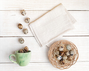Easter quail eggs on woven plate, eggs falling to green cup, cotton napkin on white wooden backdrop.