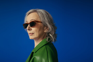 portrait of grey haired woman in green leather jacket and stylish sunglasses isolated on blue