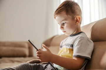 Fototapeta premium Focused little boy is sitting in a living room and playing games on a tablet.