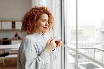 Side view indoor portrait of young attractive woman with red curly hair looking through window...