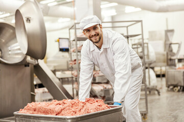 A happy meat factory worker is pushing a container full of raw ground meat in facility.
