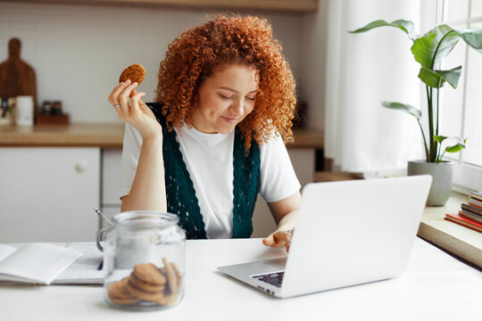 Indoor image of redhead curly young woman freelancer working on laptop sitting at kitchen table with cookie in hand and jar of cookies next to her, having coffee break. Unconscious eating