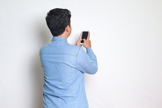 Back view portrait of excited Asian man in blue shirt holding a mobile phone and taking picture. Isolated image on white background