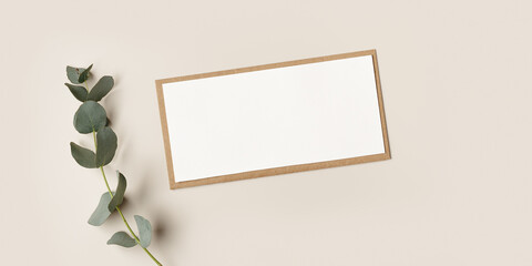 Banner, Blank paper card mock up and craft envelope with branches eucalyptus leaves, beige background. White empty invitation. Minimal modern lifestyle photo. Flat lay, top view, copy space.