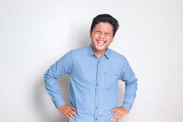 Portrait of excited Asian man in blue shirt laughing loudly while holding his chest. Fun comedy concept. Isolated image on white background