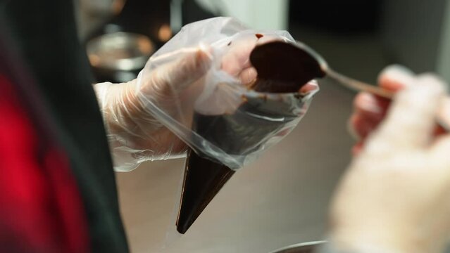 Baker putting melted chocolate into transparent pastry bag with a metal tablespoon. Blurred foreground. High quality 4k footage