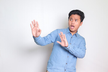 Portrait of scared Asian man in blue shirt forming a hand gesture to avoid something. Advertising...