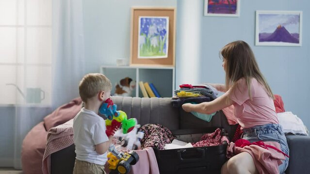 Little blond boy carries pile of toys while mother puts clothes into suitcase in living room. Preoccupied family prepares for vacation journey