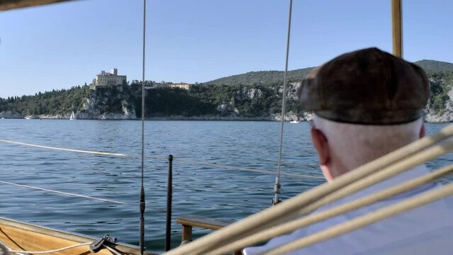man drawing on small notebook sitting on sailboat enjoying the moment with duino castle in background