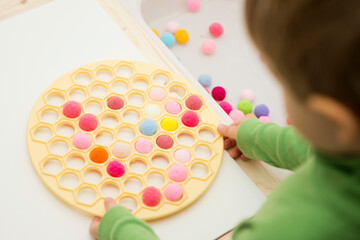 3-year-old is playing with a Russian Dumpling Maker, inserting pompoms into holes. Colored yarn balls in a plastic bag. Montessori concept. Early education, color play, fine motor skills.
