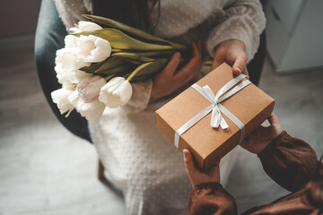 The child's hands hold a beautiful gift box with a ribbon and white tulips. Top view, close-up....