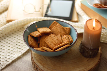 Vintage books, reading glasses, e-reader, cup of tea, bowl of biscuits and lit candle on the table. Selective focus.