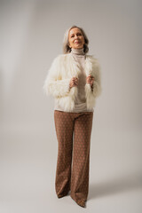 full length of pleased senior woman in faux fur jacket posing on grey background