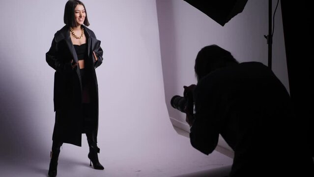 Sexy Young Fashionable Woman Model In A Black Swimsuit And Leather Boots And Coat Is Photographed In The Studio With A Photographer On A White Background. Backstage