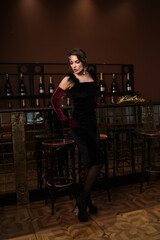 Beautiful woman at bar with a glass of vine. Bottles of vine in background. Lady portrait in black dress with make-up and hair-dress. Woman in black velvet dress and in violet gloves and black stockin