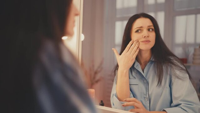 Young woman looking in mirror, unhappy with her appearance, skin imperfections