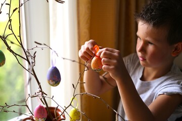 A boy of seven years of Caucasian nationality, in the kitchen at home, paints Easter eggs, decorates a branch from a tree, is engaged in creativity