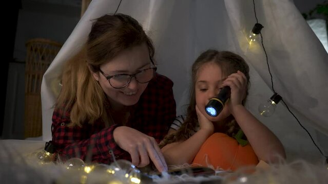 Young mother with baby is reading book with flashlight hiding in crib. Time with kids at home.smiling girl listens to mother reading book in evening before going to bed. Happy family idyll.happy baby