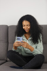 Excited black woman typing a message on smart phone. Cheerful POC female sitting on couch at home and using mobile phone for communication