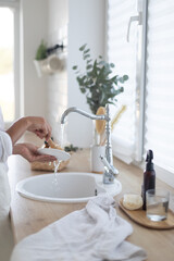 Woman washing dishes with detergent on kitchen. Eco brush with foam and basket on white bright kitchen.