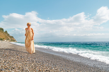 Fototapeta na wymiar Pretty blond woman walking on pebble beach enjoying sunny windy day near blue sea. Travel concept. Pastel muted color.