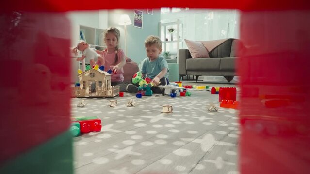 Children play with baby doll and airplane sitting near wooden house. Girl and boy spend time in nursery view through clearance in construction of cubes