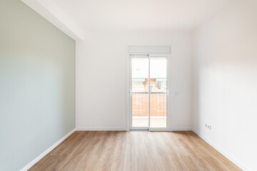An empty bright room for living with access to balcony and view of neighboring house. Room has transparent glass door with access to balcony. Gray wall and wooden laminate floor create cozy feeling.