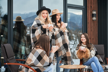 Young women enjoying winter weekends on terrace of contemporary barn house. Four girls having fun, sitting on chairs and drinking hot tea.