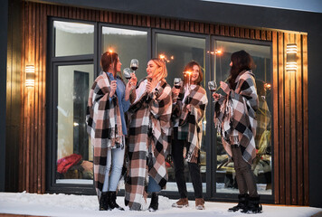 Young women enjoying winter weekends on terrace of contemporary barnhouse. Four girls in plaids drinking wine and celebrating with sparklers in the evening.