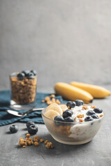 Granola cereal oatmeal with white yogurt, blueberries and banana fruits in a bowl on a blue napkin, grey background