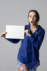 A girl in a blue shirt on a gray background holds a white sheet in her hands with a surprised face