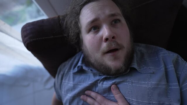 Unwell overweight man suffering from chest pains. A fat guy having heart pressure laying on sofa indoros. Close up face of male person with worried emotion