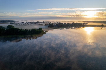 Obraz premium Foggy morning on a beautiful lake surrounded by forests and fields. A small village located near a lake in fog and morning sun.