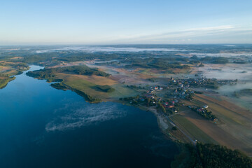 Foggy morning on a beautiful lake surrounded by forests and fields. A small village located near a lake in fog and morning sun.