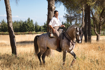 Beautiful young woman walking, riding her horse in the countryside next to several pine trees, on a sunny day. Concept horse riding, animals, dressage, horsewoman.