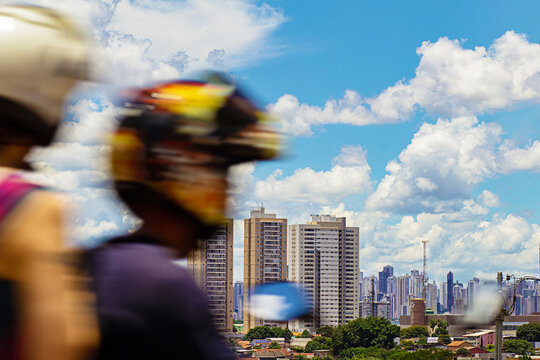 Um motociclista fora de foco e a cidade de Goi&acirc;nia vista do Morro do Al&eacute;m, com muitos pr&eacute;dios e um c&eacute;u nublado ao fundo.