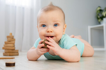 Cute little baby with big blue eyes in mint color bodysuit lying on floor at home with light background and hold pine cone in hands. Eco friendly toys. Funny baby.